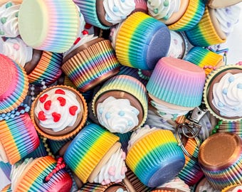 May include: A collection of colorful cupcakes with rainbow-striped wrappers. The cupcakes have chocolate frosting and are topped with white frosting and decorative sprinkles. Some have heart-shaped sprinkles. The image is a close-up shot.