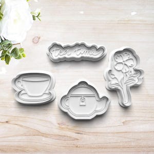 May include: A set of four white plastic cookie cutters shaped like a teapot, a teacup, a flower, and the words "tea time".