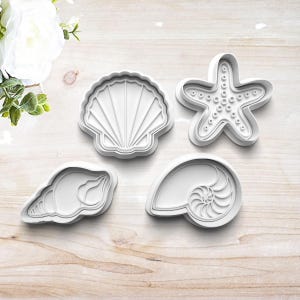 May include: Four white plastic cookie cutters shaped like seashells and a starfish. The cutters are on a light brown wooden surface.