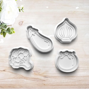 May include: A set of four white plastic cookie cutters shaped like vegetables, including an eggplant, an onion, a head of broccoli, and a tomato. Each cutter features a raised outline and internal detailing to create impressions in dough.