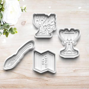 May include: Set of four white plastic cookie cutters shaped like a broom, a book, a goblet, and a tree.