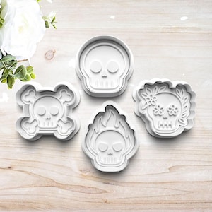 May include: Four white plastic skull-shaped cookie cutters. Designs include a classic skull, a skull with crossbones, a skull engulfed in flames, and a skull decorated with flowers. Perfect for Halloween or Day of the Dead baking.