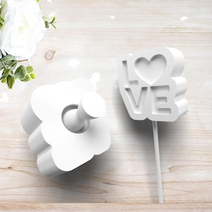 May include: Two white decorative items. One is a flower-shaped hook with a cylindrical peg. The other is a cake topper with the word "LOVE" and a heart. Both items are on a light wood surface.