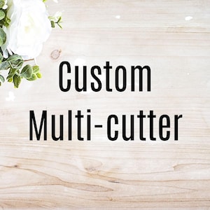 May include: The words "Custom Multi-cutter" are displayed in bold black font against a light wood grain background. White flowers and green foliage are in the upper left corner.