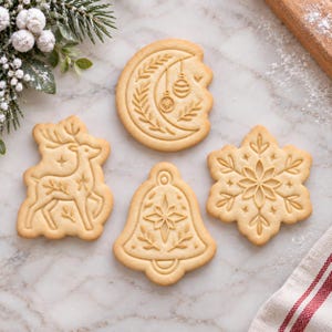 May include: Four light beige holiday cookies with embossed designs including a crescent moon with ornaments, a reindeer, a bell with a star, and a snowflake. The cookies feature intricate botanical and geometric patterns pressed into the dough.