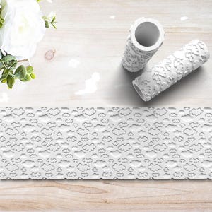 May include: White textured rolling pins with cloud and floral patterns. A strip of embossed paper with a cloud design is also visible. The items are arranged on a light wood surface with white flowers in the background.