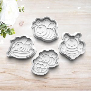 May include: A set of four white plastic cookie cutters featuring bee designs. One cutter shows a bee, another a bee on a leaf, one a bee on a flower, and the last a bee with wings spread. Each cutter has a raised edge for cutting and detailing.