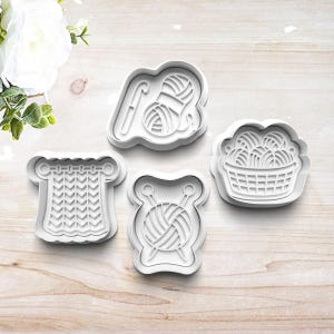 May include: Four white plastic cookie cutters with knitting and crochet themes. The shapes include a ball of yarn with a crochet hook, a knitted piece, a ball of yarn with knitting needles, and a basket filled with yarn.