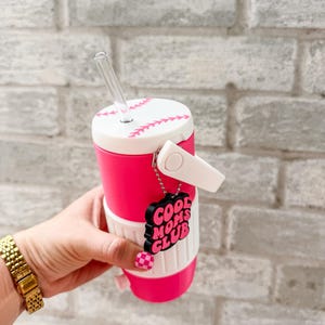 May include: A pink and white baseball-themed tumbler with a clear straw. The tumbler has a baseball stitch design and a "Cool Moms Club" charm. The tumbler is held by a person with a gold watch and pink checkered nails.