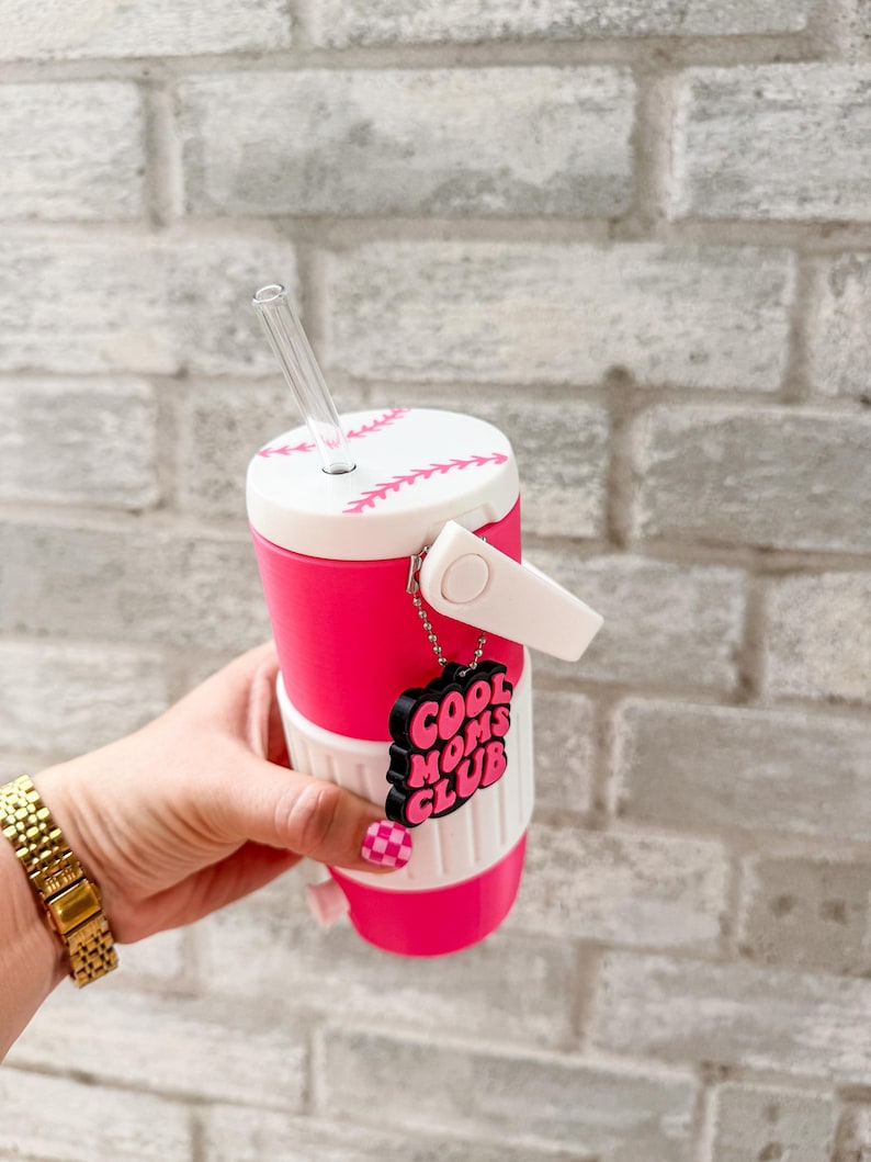 May include: A pink and white baseball-themed tumbler with a clear straw. The tumbler has a baseball stitch design and a "Cool Moms Club" charm. The tumbler is held by a person with a gold watch and pink checkered nails.