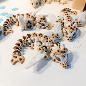 May include: Several articulated dragon figurines with white, tan, and brown details are arranged on a white surface. The dragons have cookie-like patterns and white wing accents. One dragon is in the foreground, with others in the background.