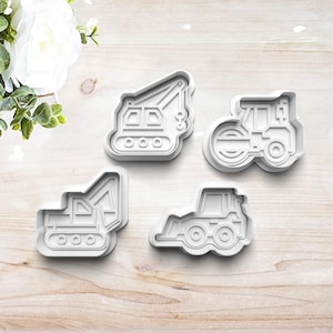May include: Set of four white plastic cookie cutters shaped like construction vehicles: a crane, a bulldozer, a backhoe, and a road roller.
