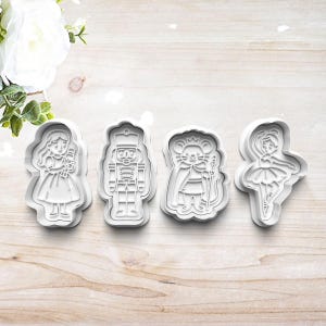 May include: Four white plastic cookie cutters in the shapes of characters from The Nutcracker ballet. The shapes include a girl, a nutcracker soldier, a mouse king, and a ballerina. The cutters are arranged on a light wood surface.