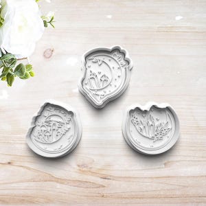 May include: Three white plastic cookie cutters with moon and crystal designs. The cutters feature a crescent moon with stars, a crescent moon with crystals and flowers, and a crescent moon with mushrooms and a dragonfly.