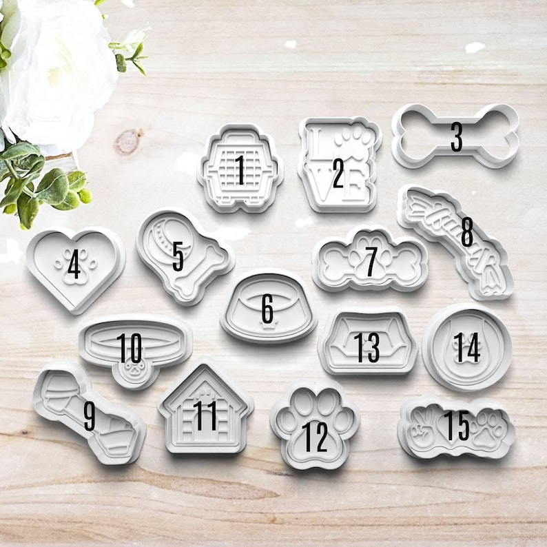 May include: A set of 15 white plastic cookie cutters shaped like various dog-related items, including a bone, a dog house, a paw print, a collar, a bowl, and a heart. Each cutter is numbered 1 through 15.