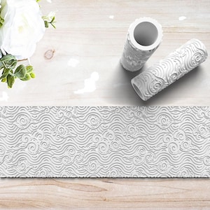 May include: Two white textured pattern rollers with a matching embossed pattern. The rollers are cylindrical with a hollow center. A strip of the embossed pattern is displayed below the rollers. The pattern features swirling wave designs.