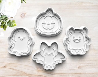 May include: Four white Halloween-themed cookie cutters. The shapes include a smiling ghost, a jack-o'-lantern, a bat, and a monster. The cutters are arranged on a light wood-grain surface. Perfect for baking seasonal treats.