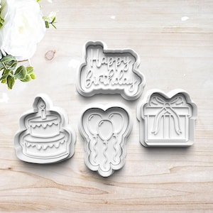 May include: Four white plastic cookie cutters in the shapes of a birthday cake, balloons, a gift box, and the words "Happy Birthday". The cutters are arranged on a light wood surface.