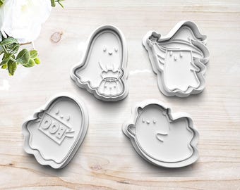 May include: Four white plastic Halloween-themed cookie cutters. Designs include a ghost holding a pumpkin bucket, a ghost wearing a witch hat, a ghost with a "BOO" tombstone, and a smiling ghost. The cutters are on a light wood surface.