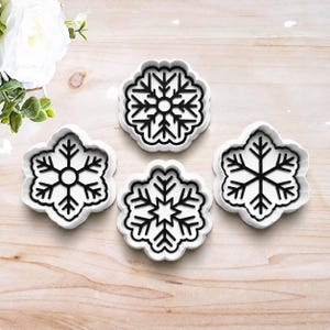 May include: A set of four white plastic cookie cutters, each featuring a distinct black snowflake design. The cutters have a scalloped outer edge and a detailed internal structure for creating snowflake-shaped cookies with intricate patterns.