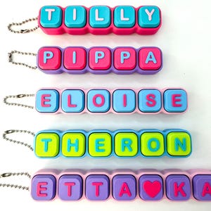 May include: Keychains with names spelled out in colorful letter blocks. The names Tilly, Pippa, Eloise, Theron, and Etta are displayed. Each keychain has a different color scheme, with a silver chain attached.