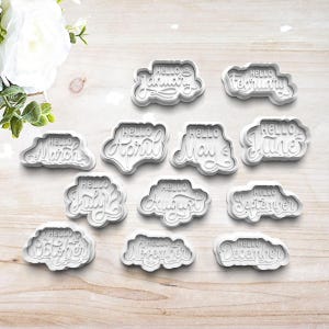 May include: A collection of white plastic cookie cutters, each shaped like a cloud and bearing the word "Hello" followed by a month of the year. The months include January, February, March, April, May, June, July, August, September, October, November, and December.
