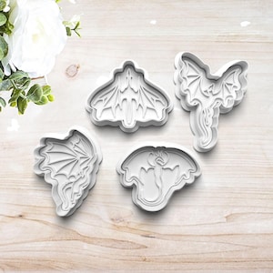 May include: Four white plastic cookie cutters shaped like dragons. The cutters are on a light brown wooden surface.