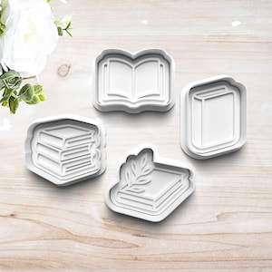 May include: Four white plastic cookie cutters in the shape of books. One cutter is an open book, another is a closed book, and two are stacks of books. One stack of books has a leaf design.