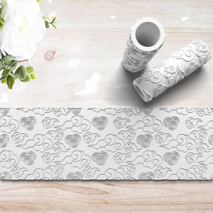 May include: Two white textured pattern rollers with a cloud design, alongside a matching embossed pattern. The rollers are cylindrical, and the pattern is repeated on the embossed surface. The design features swirling clouds and circular motifs.