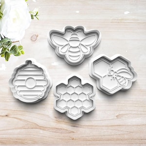 May include: Four white plastic cookie cutters in bee and honeycomb shapes. The cutters include a bee, a honeycomb, a beehive, and a bee on a honeycomb. The cutters are on a light wood surface.