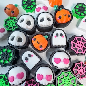 May include: A collection of Halloween-themed charms featuring pumpkins, ghosts, skulls, and spiderwebs. The charms are in various colors, including orange, white, black, green, and pink. The charms are likely for crafting or jewelry making.