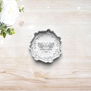 May include: White plastic cookie cutter shaped like a wreath with a bee in the center.