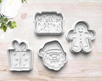 May include: Four white plastic Christmas-themed cookie cutters. Designs include a gift box, Santa Claus face, gingerbread man, and a rectangular cutter with the words "MERRY CHRISTMAS".