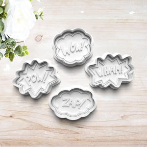 May include: Four white plastic cookie cutters shaped like speech bubbles with the words "POW!" "WHAM!" "ZAP!" and "WOW!" inside.