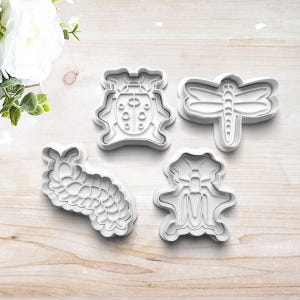 May include: Four white plastic cookie cutters in the shapes of insects. The set includes a ladybug, dragonfly, caterpillar, and a bee. The cutters are on a light wood-grain surface, with a white flower in the background.