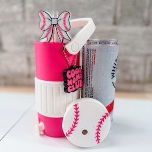 May include: A pink and white baseball-themed can cooler with a matching lid and a bow-shaped straw topper. The cooler has a handle and a charm that says "COOL MOMS CLUB". A can of White Claw is next to the cooler.