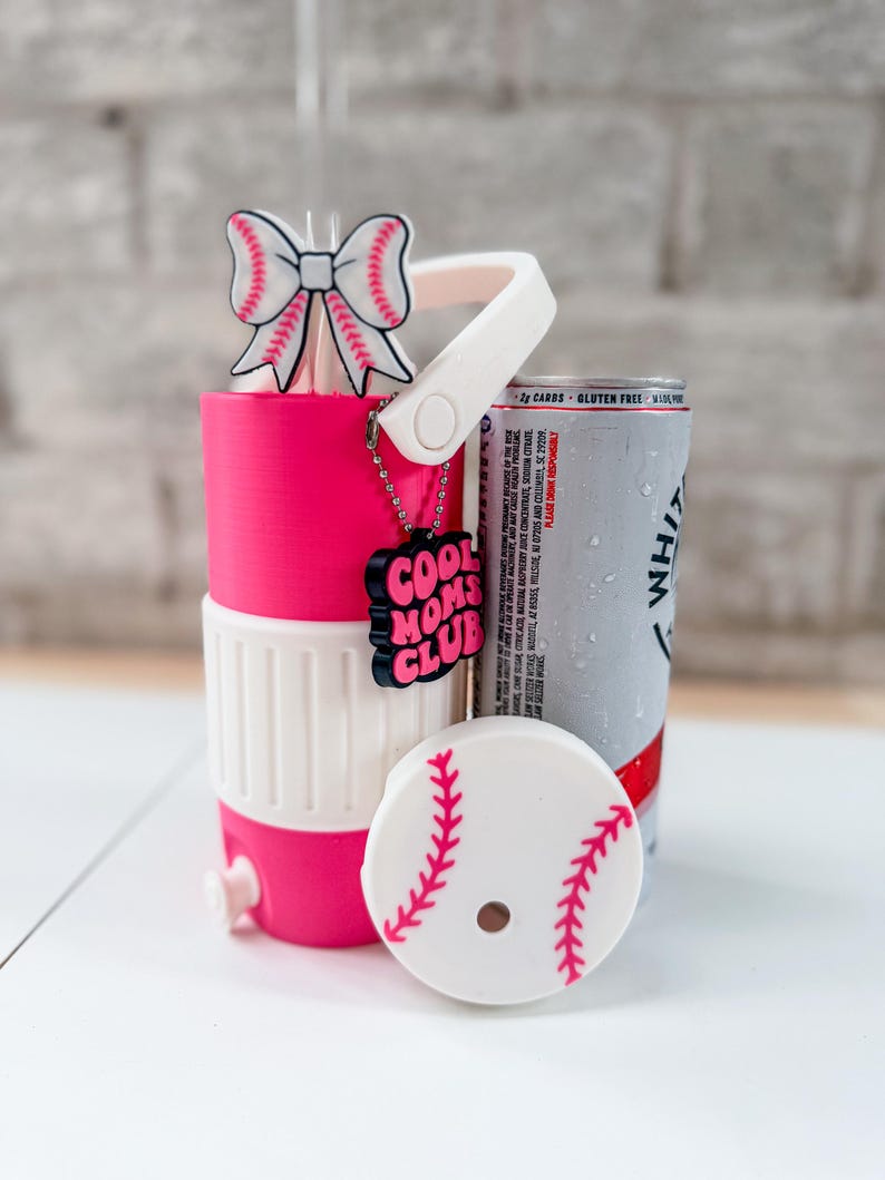 May include: A pink and white baseball-themed can cooler with a matching lid and a bow-shaped straw topper. The cooler has a handle and a charm that says "COOL MOMS CLUB". A can of White Claw is next to the cooler.