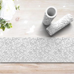 May include: Two white cylindrical patterned rollers featuring embossed Halloween motifs including skulls, ghosts, pumpkins, spider webs, bats, and the word boo. The rollers have a hollow center and a textured surface designed for creating repeating patterns.