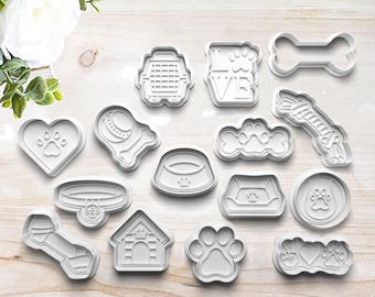 May include: A set of 15 white plastic dog-themed cookie cutters. The cutters include a bone, a dog house, a dog bowl, a paw print, a dog collar, a dog leash, a dog toy, a heart with a paw print, a dog bed, a dog cage, a dog tag, and a dog silhouette.