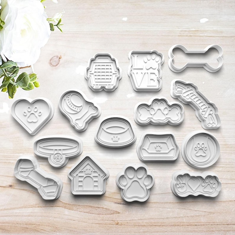 May include: A set of 15 white plastic dog-themed cookie cutters. The cutters include a bone, a dog house, a dog bowl, a paw print, a dog collar, a dog leash, a dog toy, a heart with a paw print, a dog bed, a dog cage, a dog tag, and a dog silhouette.