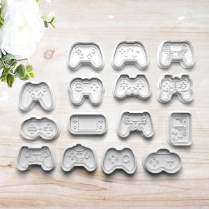 May include: A collection of white plastic cookie cutters in various video game controller shapes. The cutters include designs of classic and modern game controllers, as well as a handheld gaming device. The cutters are arranged on a light wood surface.