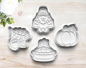 May include: Four white plastic cookie cutters in Thanksgiving shapes: a turkey wearing a pilgrim hat, a pumpkin, a pie, and an acorn with oak leaves. The cutters are arranged on a light wood surface.