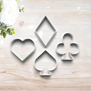 May include: A set of four white plastic cookie cutters shaped like playing card suits, including a heart, a diamond, a spade, and a club. Each cutter features a clean, open-frame design with defined edges for cutting dough into distinct shapes.