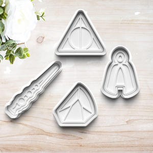 May include: Four white plastic cookie cutters shaped like a wand, a diamond, a wizard in a cloak, and the Deathly Hallows symbol.
