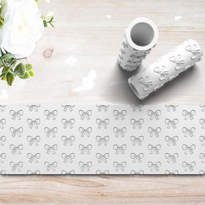 May include: Two white, cylindrical embossing rollers with a bow pattern, alongside a sheet of embossed paper with the same design. The rollers are on a light wood surface, with white flowers in the background.
