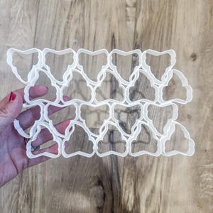 May include: A collection of white plastic cookie cutters in a repeating pattern, held against a wooden surface. The cutters have a decorative shape, possibly for creating unique cookies or pastries. The design is intricate and detailed.