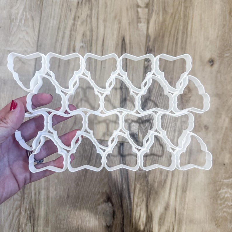 May include: A collection of white plastic cookie cutters in a repeating pattern, held against a wooden surface. The cutters have a decorative shape, possibly for creating unique cookies or pastries. The design is intricate and detailed.