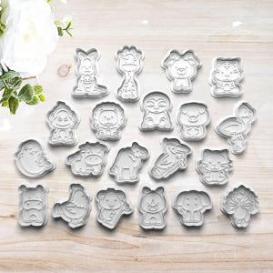 May include: A collection of white plastic cookie cutters in various animal shapes, including a kangaroo, giraffe, lion, and elephant. The cutters are arranged on a light-colored wooden surface, perfect for baking themed cookies.