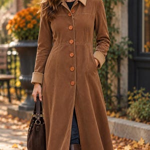 May include: A long brown corduroy coat featuring a structured collar, a front button closure with six round buttons, long sleeves with ribbed cuffs, and two side pockets. The coat has a tailored waistline and a flared skirt that reaches below the knee.