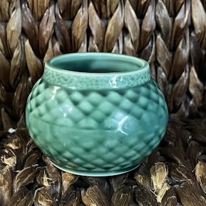 May include: A small, round, light green ceramic bowl featuring a textured, diamond-shaped lattice pattern across its exterior surface. The bowl has a smooth, glossy finish and a slightly tapered rim with a subtle, dotted border detail.